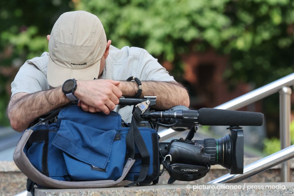 A cameraman is leaning over his camera which is in the foreground. His face is covered by a baseball cap.