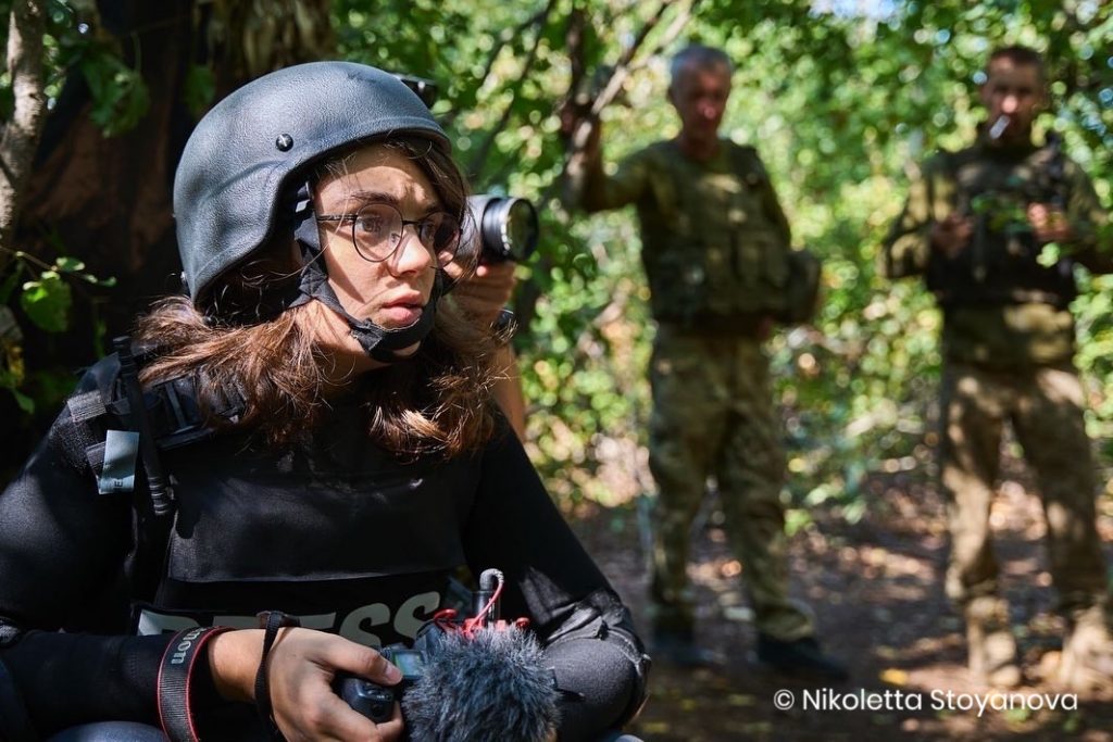 Eine Journalistin mit Helm und Schutzweste hält eine Kamera in ihrer Hand. Im Hintergrund stehen zwei Personen in Armeebekleidung.
