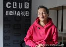 Eine Frau sitzt in einem pinken Kapuzenpullover vor einer schwarzen Wand, auf der "Club Belarus" zu lesen ist.