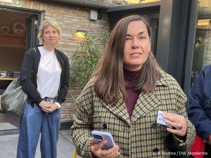 Eine Frau in einem Mantel hält ihr Smartphone in der rechten Hand, während im Hintergrund eine weitere Frau steht