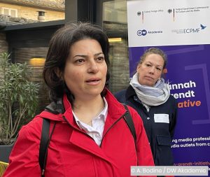 Eine Frau in einer roten Jacke spricht, während eine Zuhörerin im Hintergrund steht